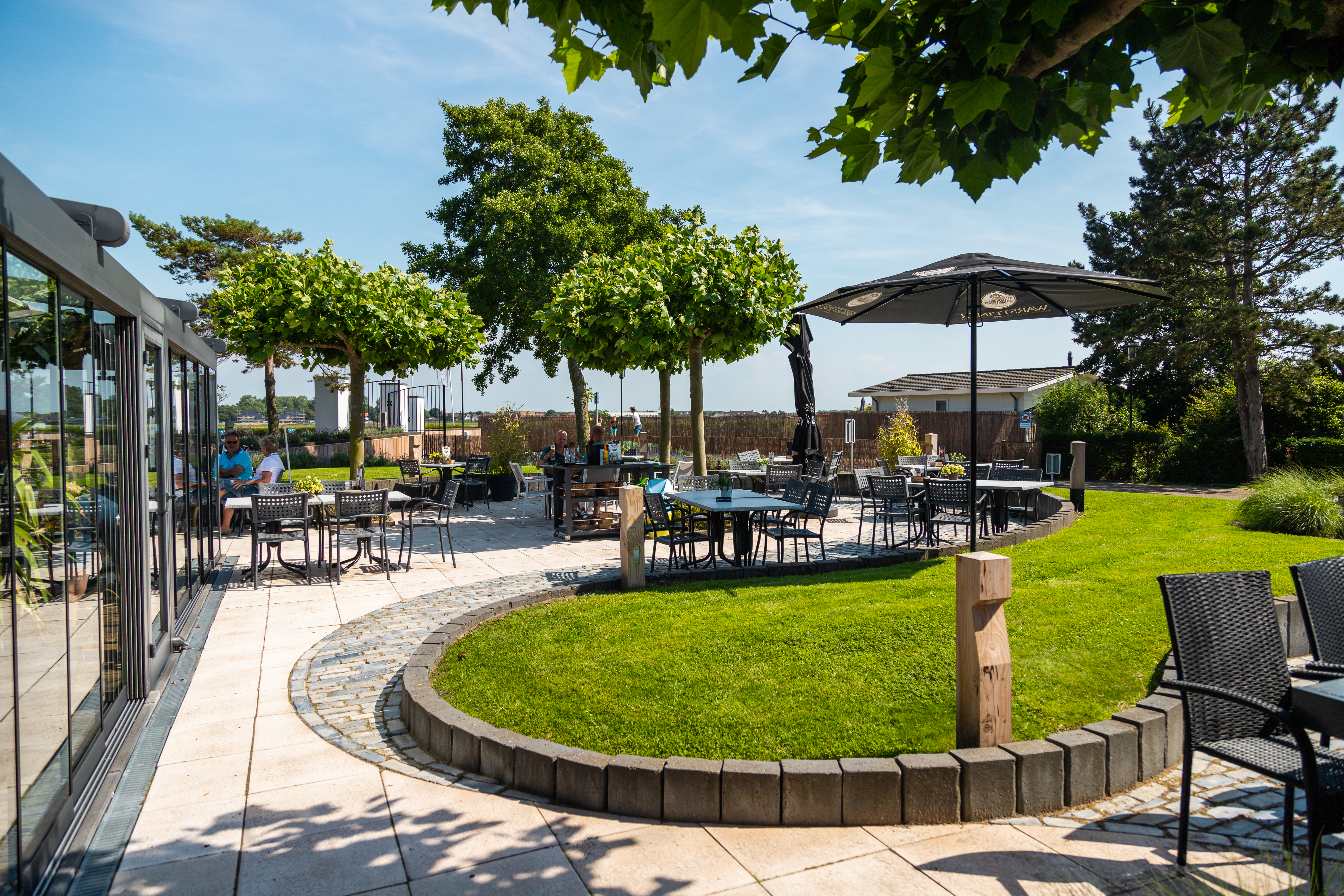 Courtyard tables under trees, people dining and conversing, with a lush, green lawn and clear blue sky at Europarcs Parc du Soleil