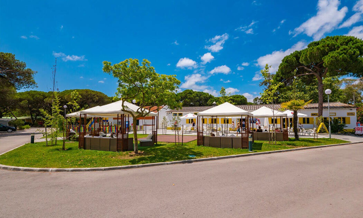 White canopies provide shade over outdoor seating on a sunny day, surrounded by trees and green grass at Costa da Caparica