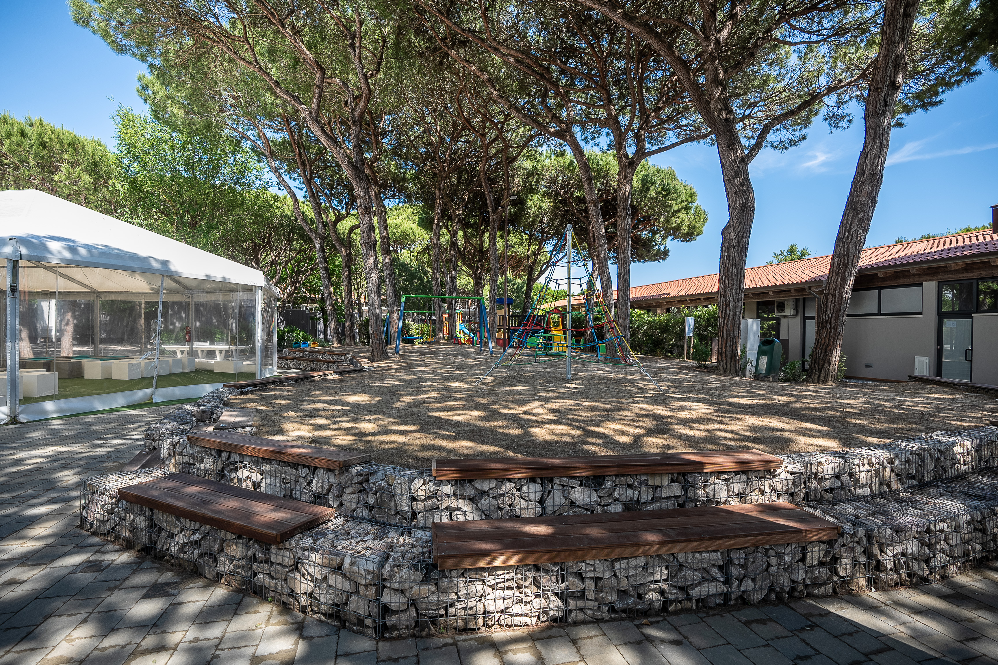 Playground with climbing structure and swings under tall trees, next to a white tent and building at Malibu Beach