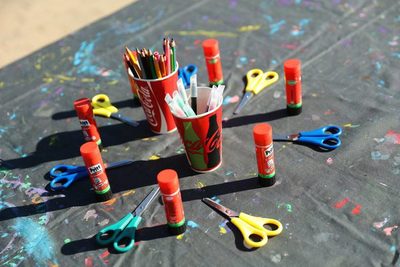 Colorful craft supplies, including scissors, glue sticks, and pencils, arranged on a paint-splattered table outdoors at Petite Suisse