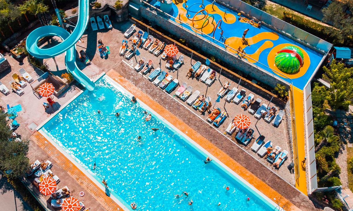 A swimming pool with people, surrounded by lounge chairs and umbrellas, next to a colorful water park play area at Eden