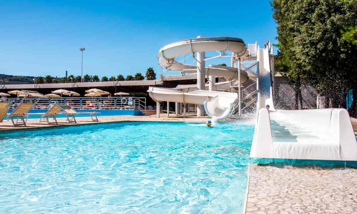 Curved waterslide empties into a busy outdoor pool surrounded by lounge chairs, umbrellas, and trees at Villaggio Turistico Lido D Abruzzo