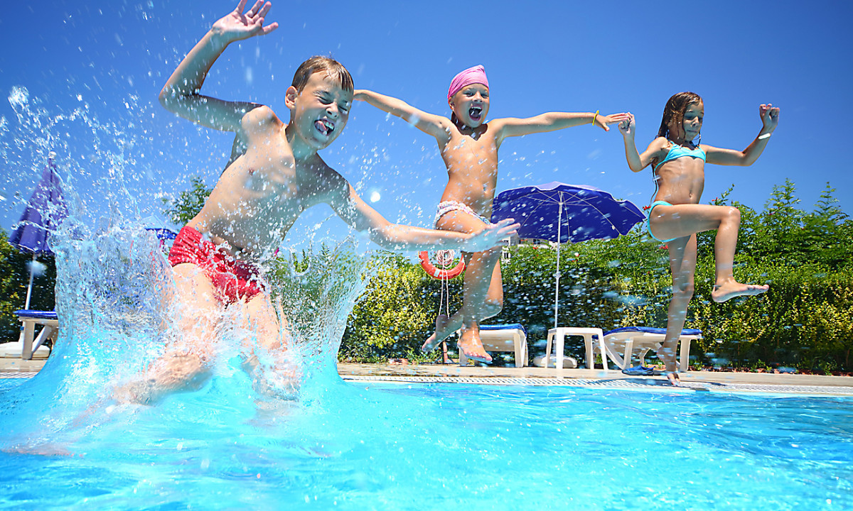 Three children are joyfully jumping into a swimming pool, splashing water around with lounge chairs and umbrellas in the background at Mar Y Sierra