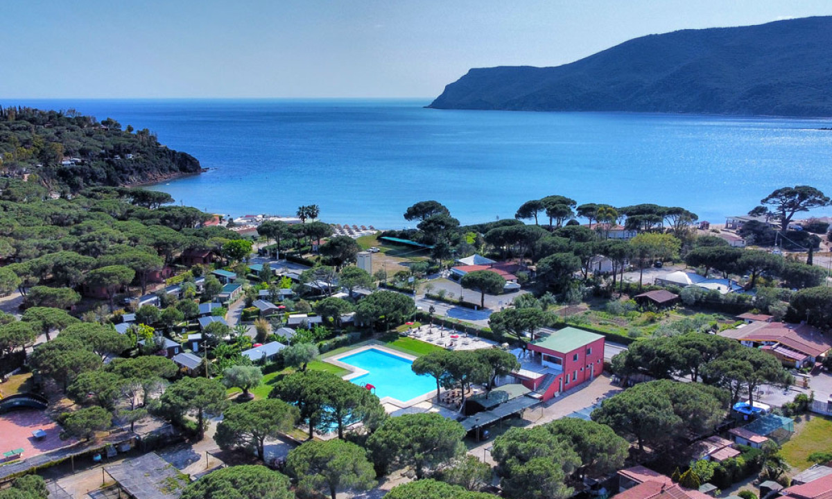Coastal campsite features a vibrant pool area surrounded by lush trees, adjacent to a calm, expansive sea at Camping Lacona Pineta