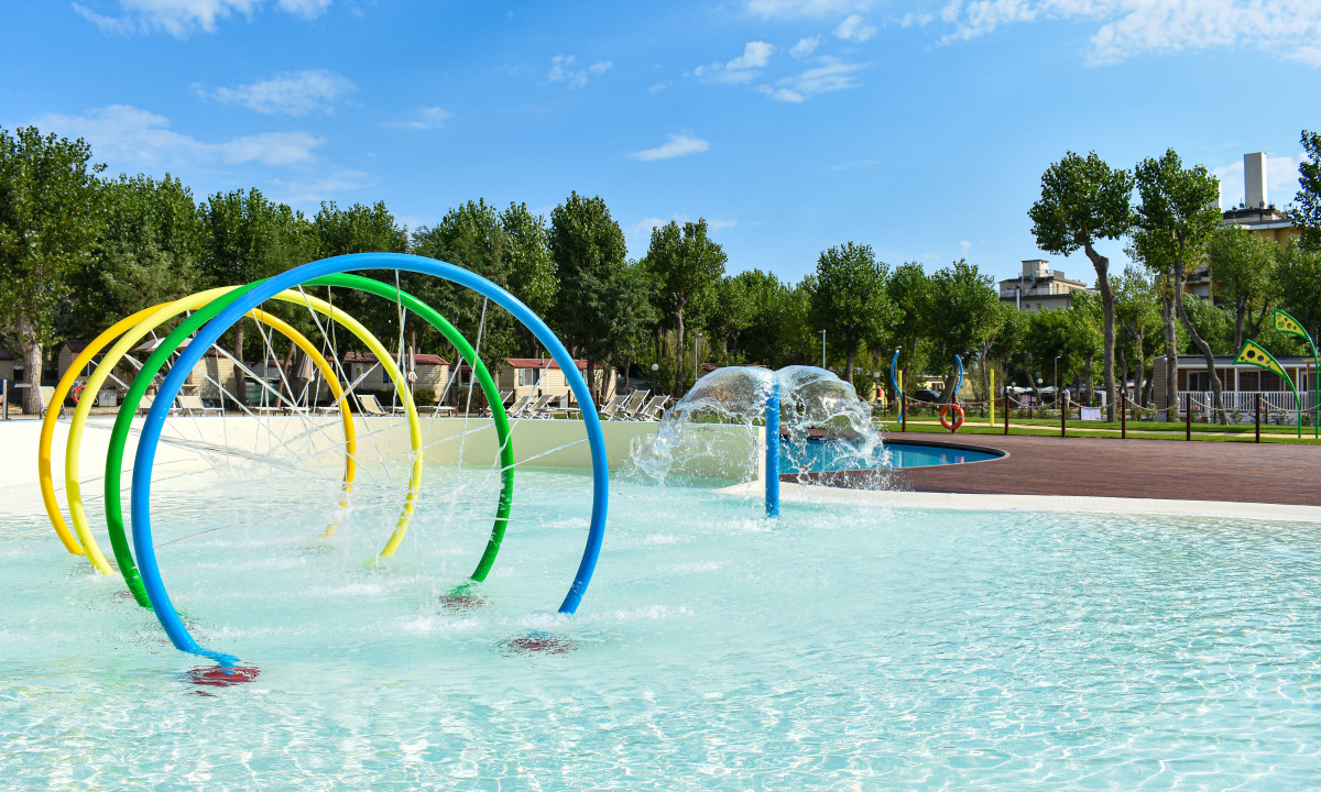 Colorful water spray arches creating a playful mist in a shallow pool, surrounded by trees and recreational facilities at Rimini Family Camping Village